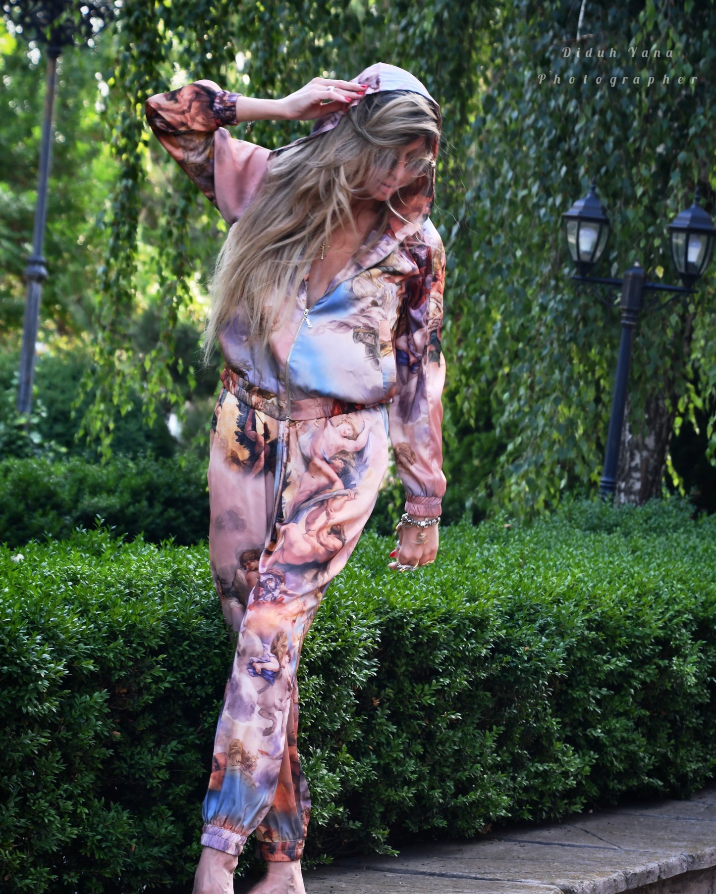 A person wearing a multicolored jumpsuit with a floral and abstract pattern, striking a playful pose on one leg on a stone bench with greenery in the background.