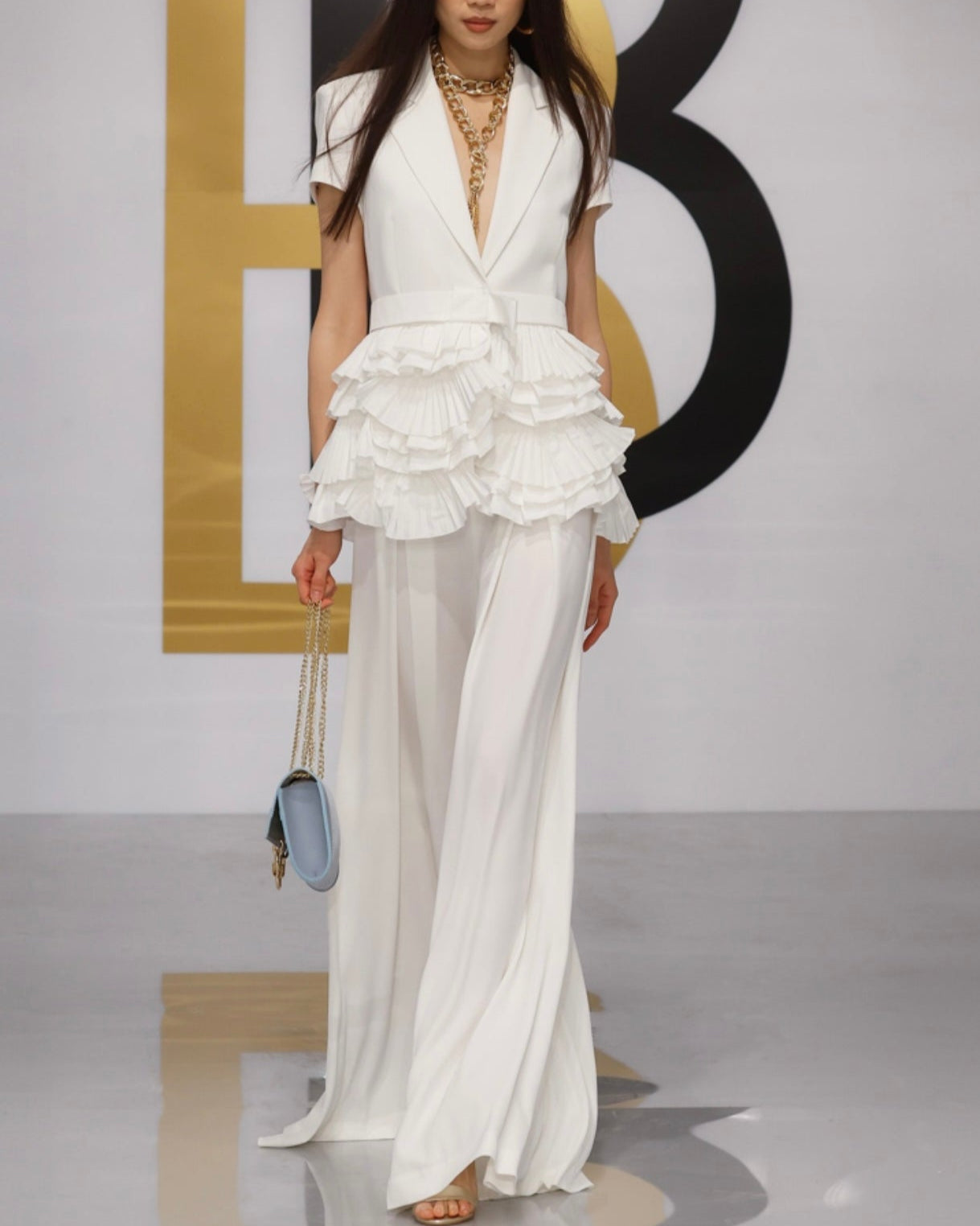 A model walking a fashion runway wearing a white jacket with frills and a matching white skirt, accessorized with a necklace and carrying a handbag.