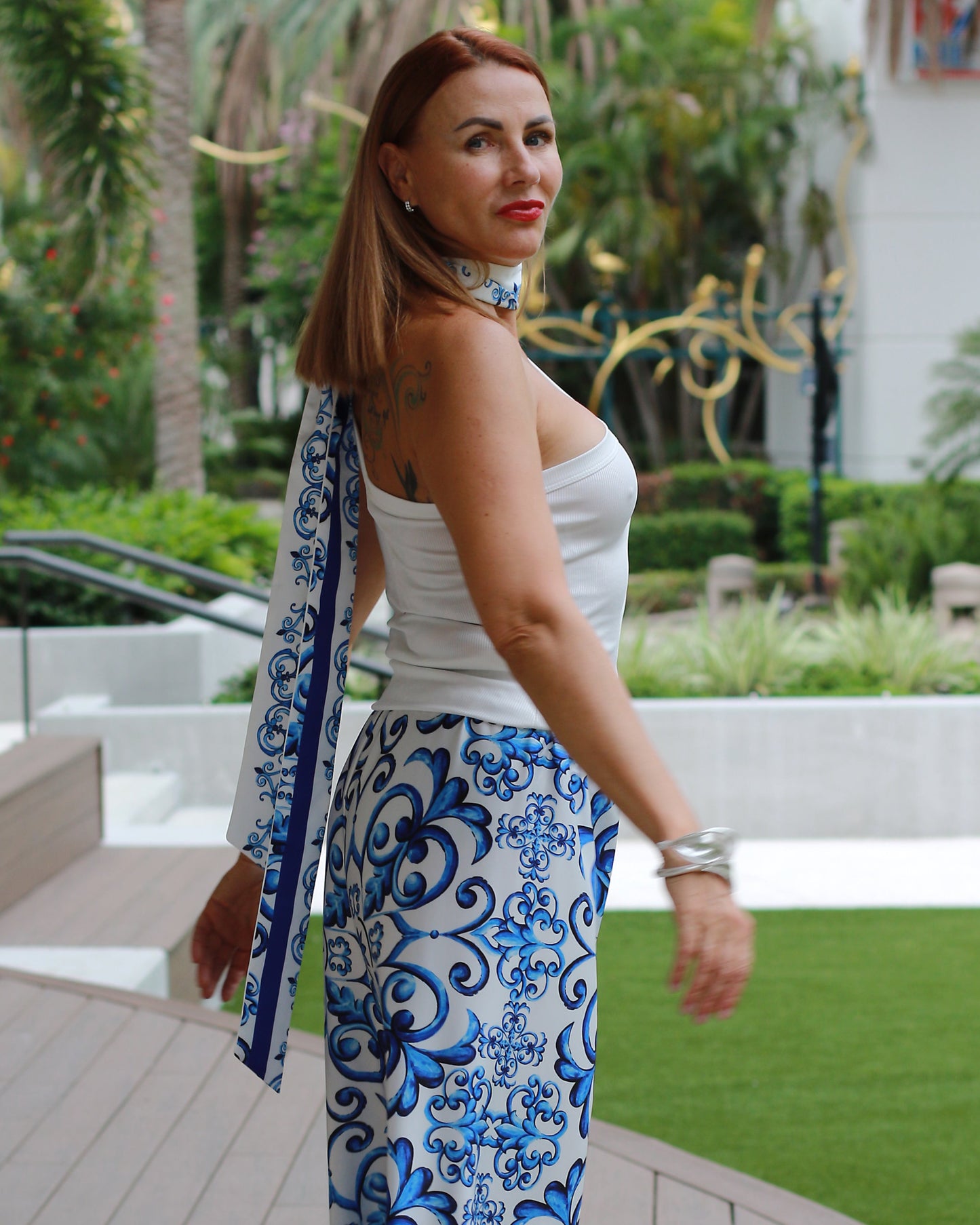 Woman in a white top and blue patterned pants standing on a wooden deck with greenery in the background
