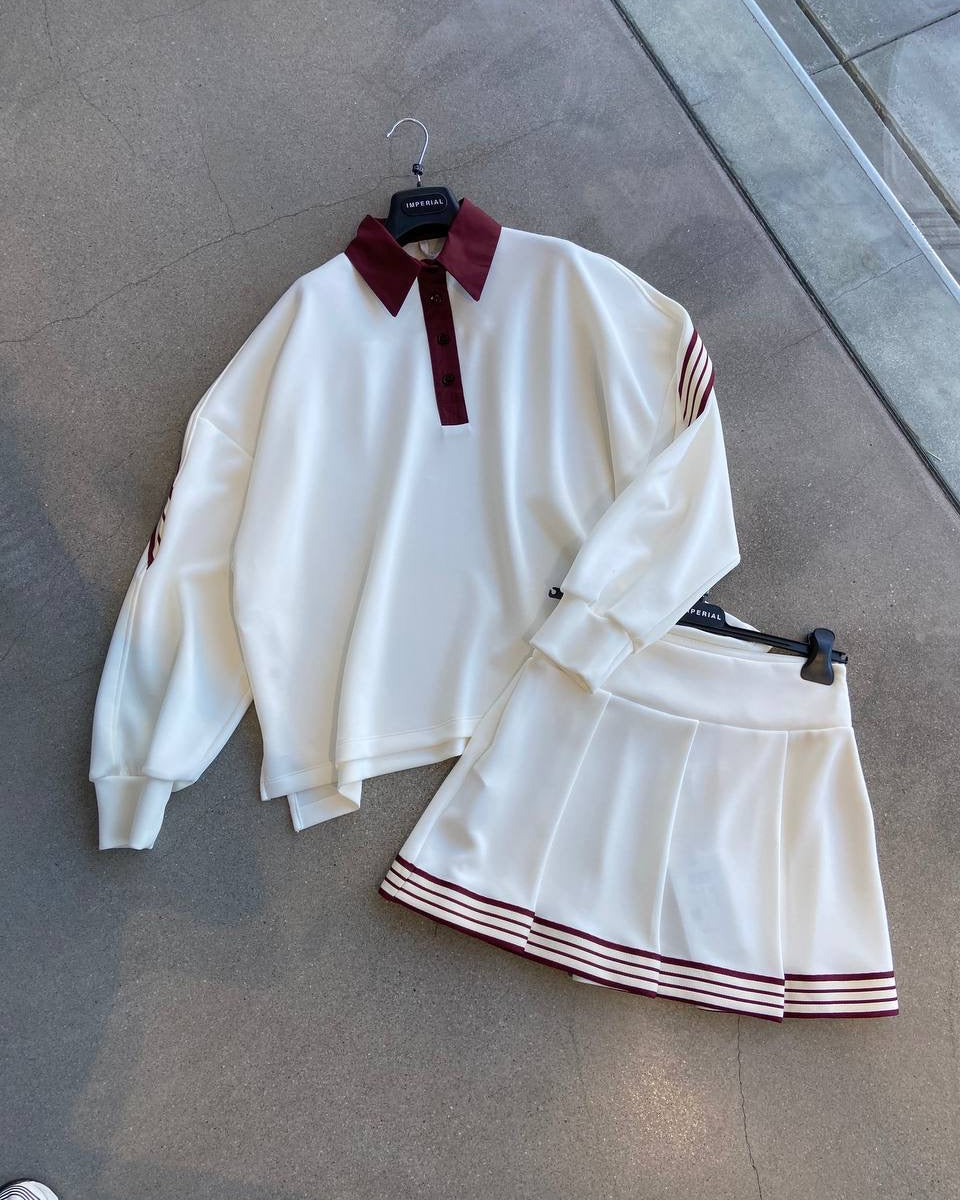 A beige golf skirt with red stripes and a matching long-sleeved golf shirt with a collar, displayed on hangers against an indoor background.
