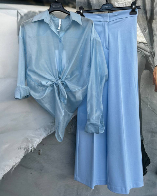 A blue blouse tied at the waist and displayed next to matching blue wide-leg trousers on hangers against a concrete backdrop.