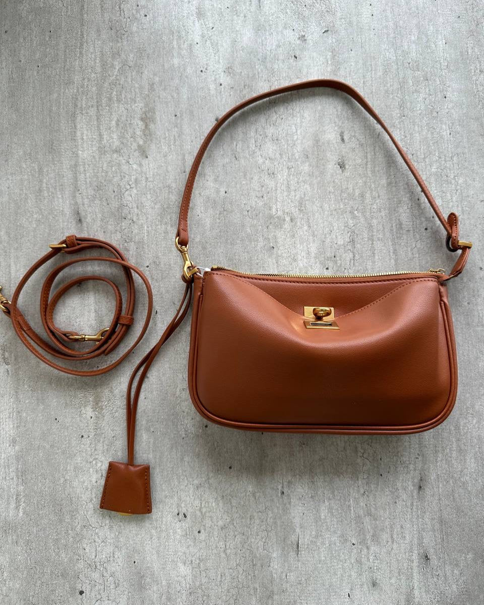 A brown leather handbag with a shoulder strap and a front lock closure, placed on a concrete surface.