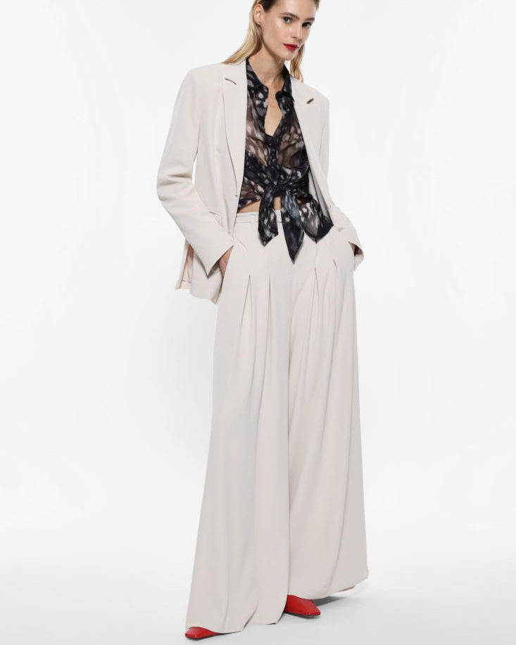 A model wearing a beige wide-leg pants and a black floral blouse with a tie-neck