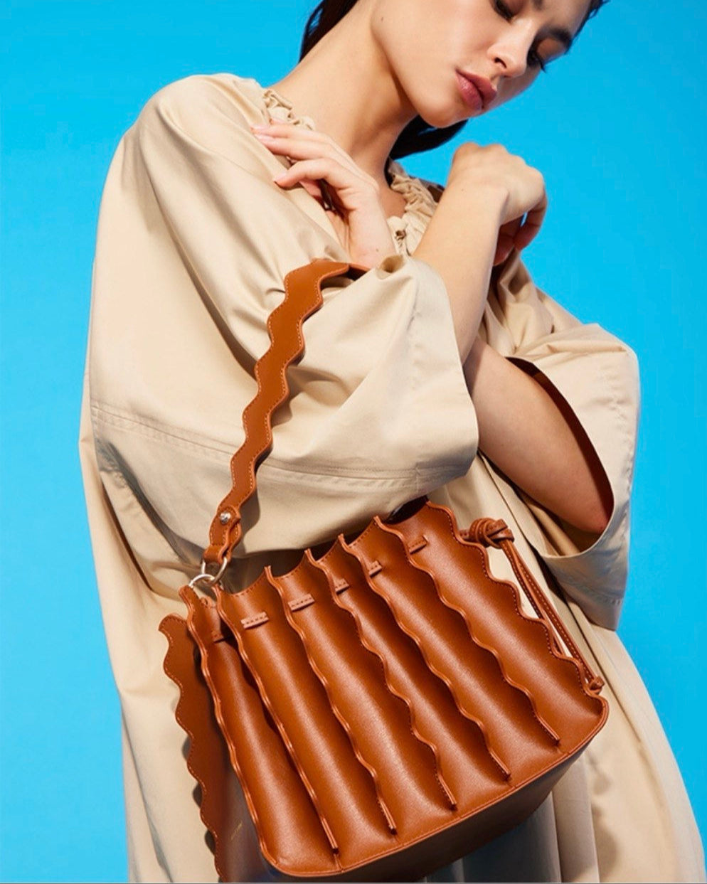 A person wearing a beige coat and holding a brown bucket-shaped handbag with a textured surface.