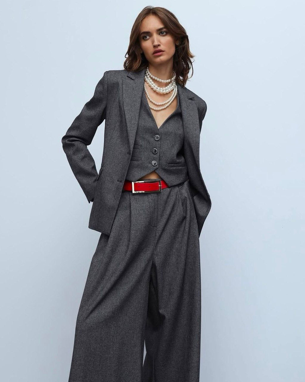 A person wearing a gray suit with a red belt, consisting of a jacket and trousers. Accessory details include necklaces and a watch.