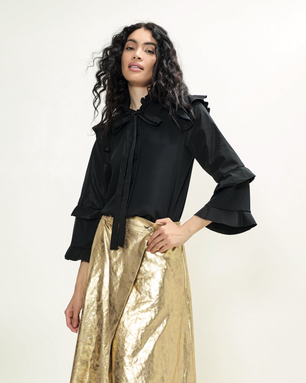 A woman modeling a black blouse with ruffled sleeves and a round neckline, paired with a metallic gold skirt. She is wearing black boots and standing against a white background.