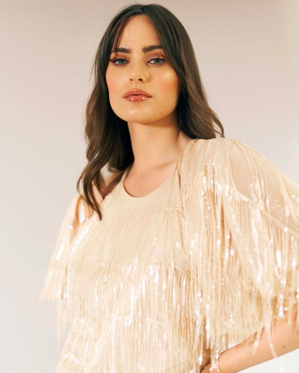 A woman wearing a beige dress with sequins and fringes across the bottom, standing against a neutral background.