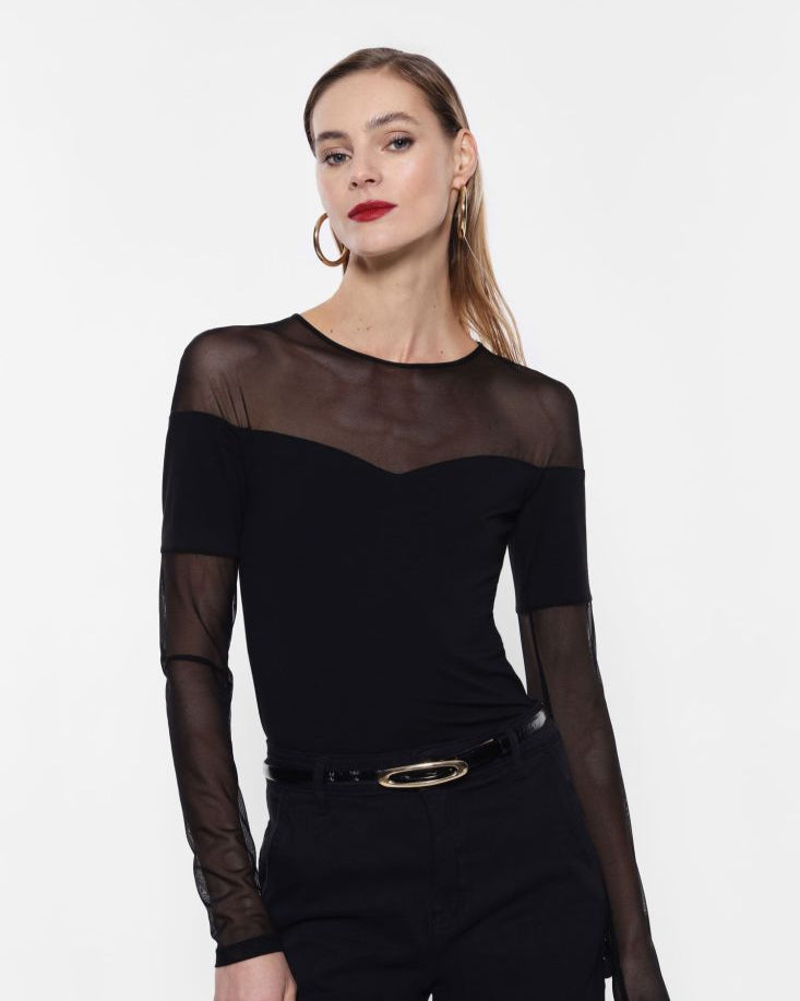 A woman wearing a black long-sleeved top with a sheer mesh neckline, paired with black trousers. Accessories include hoop earrings and a belt.