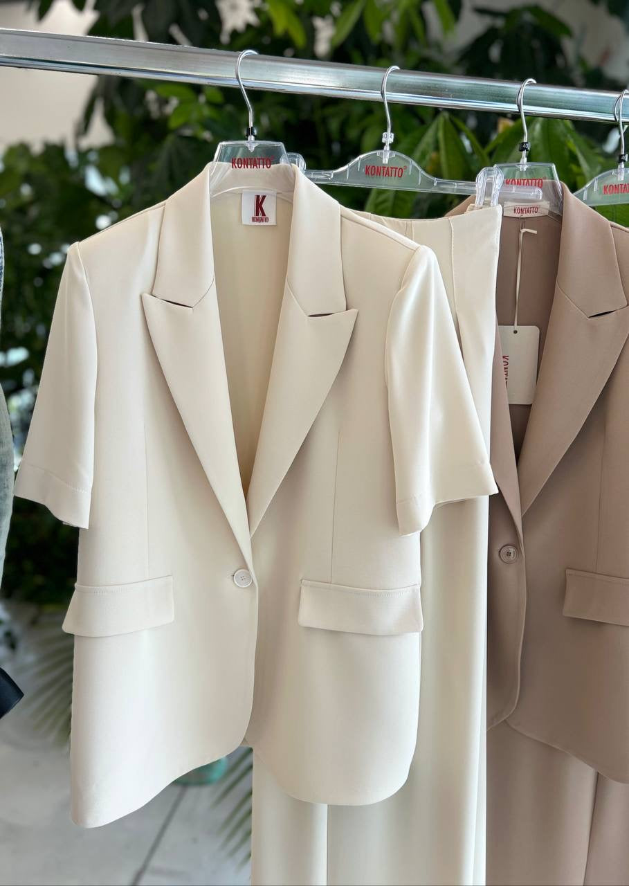 Beige blazer and skirt set on hangers with tags against a blurred green background