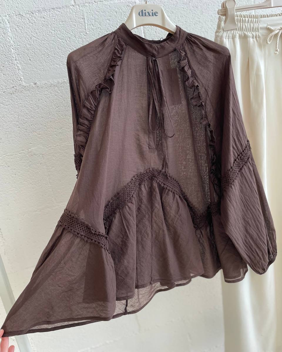 Brown blouse with ruffles on a hanger against a white textured wall.