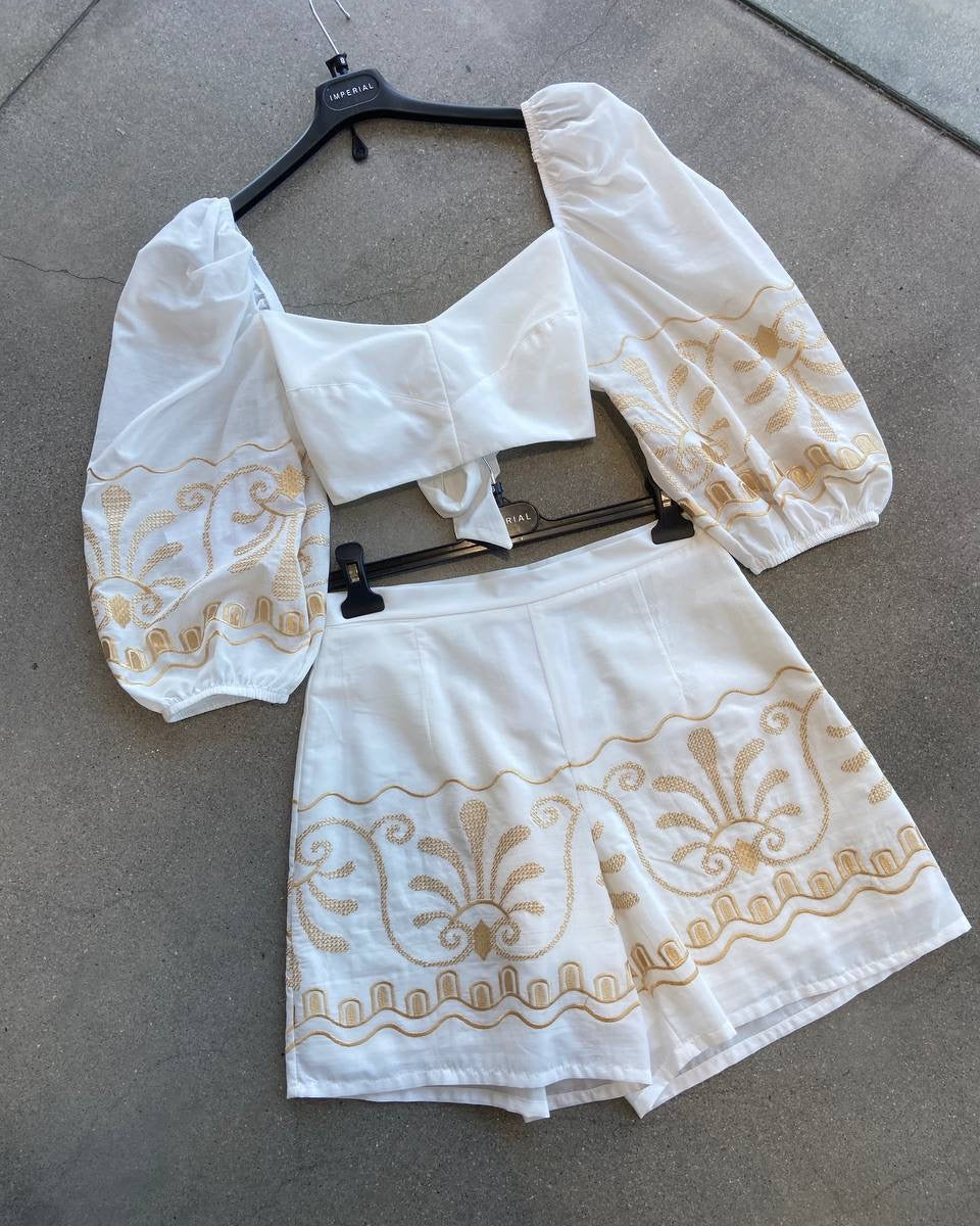 White blouse and shorts with gold embroidery on a hanger against a gray background