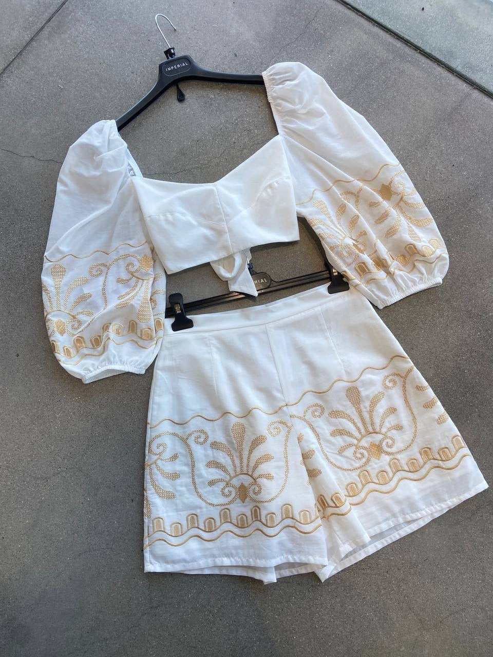 White blouse and shorts with gold embroidery on a hanger against a gray background