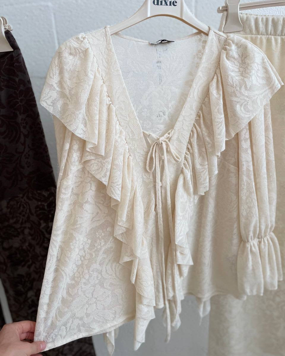 White lace blouse on a hanger against a white brick wall.