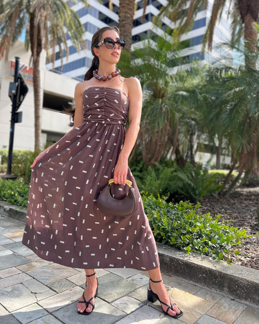 Woman in a brown strapless dress with white patterns standing outdoors with palm trees and buildings in the background.