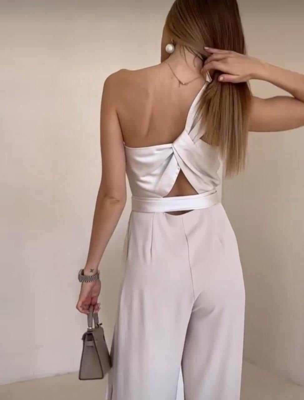 Woman wearing a white jumpsuit with cut-out design, holding a handbag against a plain background.