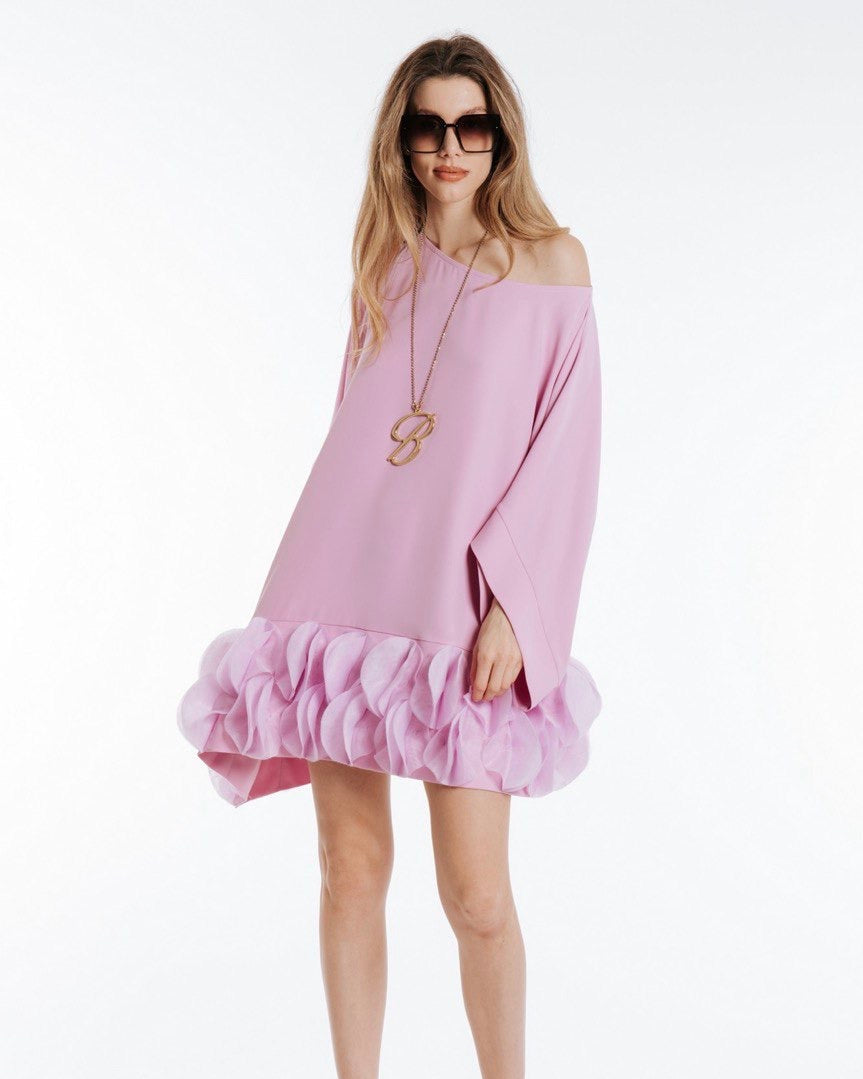 A woman standing, wearing a pink dress with a tulle ruffled hem, off-shoulder top, and paired with white sandals. She has accessorized with a long necklace and is posing with her hands at her sides.