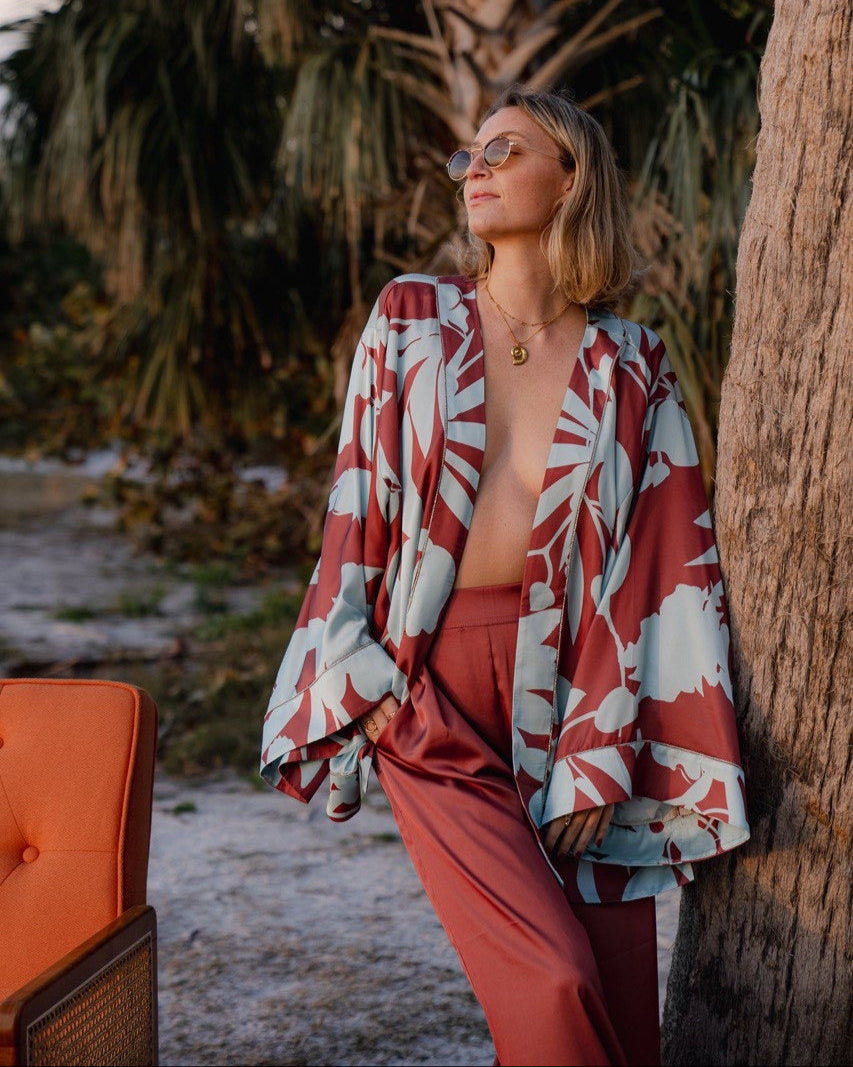 A person wearing a multicolored floral kimono with bell sleeves and matching pants, sitting next to a palm tree.
