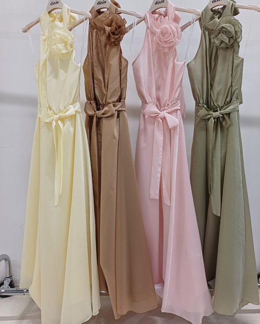 A selection of four sleeveless chiffon maxi dresses with rosette detail and bow back in different colors, displayed on hangers.