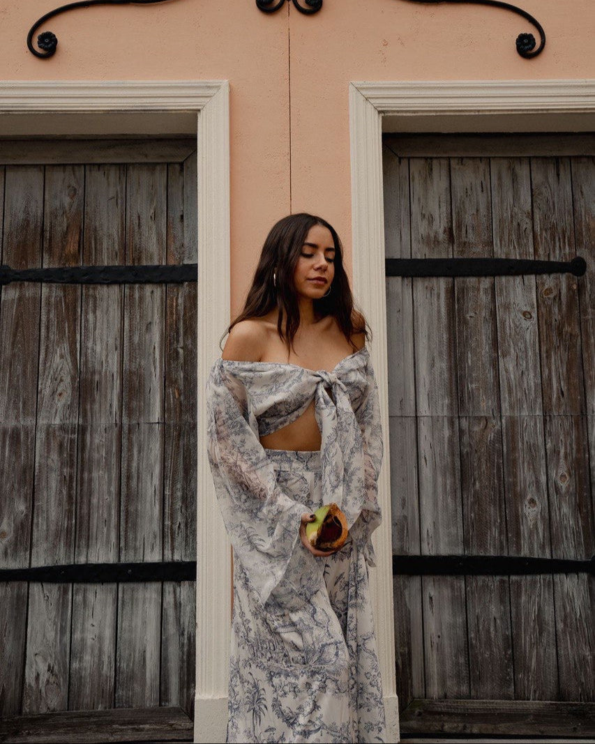 A woman standing in front of a building with wooden shutters, wearing a gray and white floral chiffon dress with long sleeves and a tie at the waist.