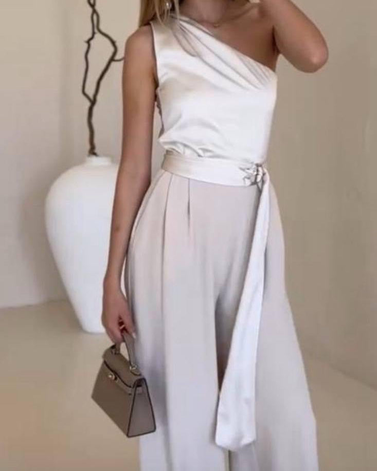 Woman wearing a white one-shoulder dress with a belt, holding a small handbag against a neutral background.
