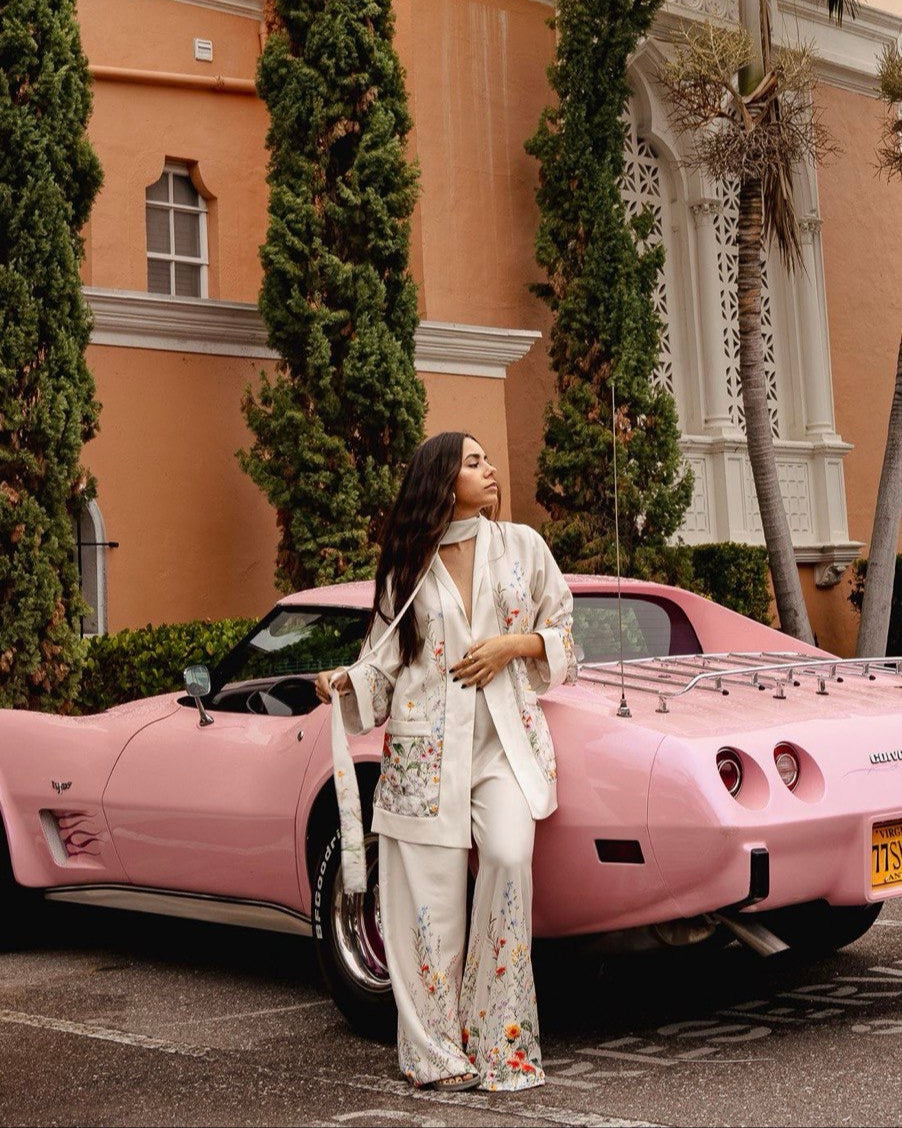 A person standing next to a pink car, wearing a white suit with a pattern.